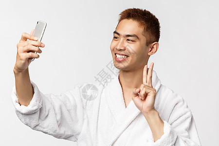 美容 水疗和技术概念 穿着浴袍的热心亚洲男子在酒店休息时自拍 在 covid19 期间与家保持距离 展示和平标志并在手机上拍照图片