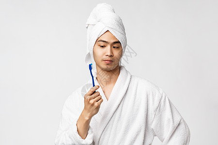 美容 水疗和休闲概念 穿着浴袍和浴巾的好奇而体贴的亚洲男人的画像 看着牙刷很感兴趣 思考 站在白色背景上思考图片