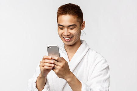 美容 水疗和技术概念 穿着浴袍的帅气亚洲男人看着智能手机显示屏很高兴 给度假酒店度假村的朋友发短信 站在白色背景下拿着手机图片