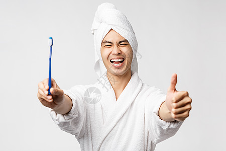 美丽 温泉和休闲概念 穿着浴袍和浴巾的热情英俊亚洲男子肖像 展示满脸满意面孔的牙刷 在家具中做拇指 推荐保健产品图片