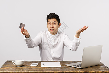 我不知道该买什么 困惑 不确定和困惑的中国男性坐在办公室里 靠近笔记本电脑 文件 拿着信用卡 耸耸肩试图决定网上的订单 白色背景图片