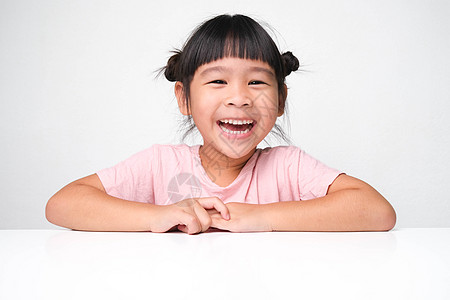 可爱的黑发女孩开心地笑着坐在一张粉红色背景的桌子旁 看着镜头 广告儿童用品图片