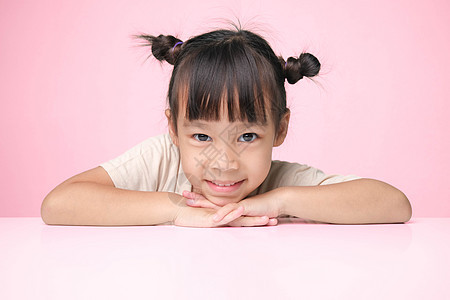可爱的黑发小女孩开心地笑着 把手放在下巴下 坐在粉红色背景的桌子旁 看着镜头 广告儿童用品图片