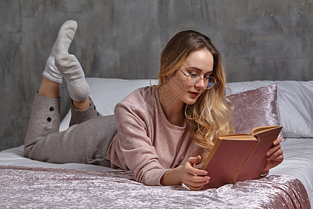 穿着眼镜 散装的金发女性 她躺在床上和卧室阅读书上 学生 博客 内墙灰色 贴近图片