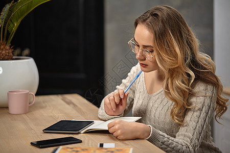 戴眼镜的模范 米色毛衣 坐在桌子上 用杯子和手掌放在桌上;在笔记本中写一些东西图片