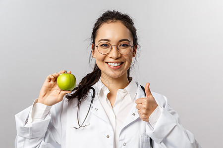 年轻开朗的亚洲女医生 治疗师或糖尿病专家的画像谈论健康饮食 展示青苹果和竖起大拇指 建议吃水果 微笑鼓励照顾健康治疗维生素暴发流图片