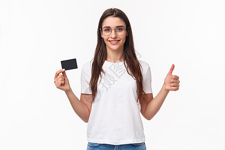 自信快乐 微笑的年轻女性穿着 T 恤 眼镜 竖起大拇指并点头表示赞同 推荐银行 出示信用卡 使用非现金支付在线购买电话金融卡片女图片