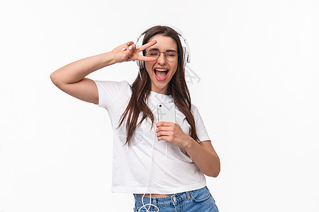 技术 生活方式和音乐概念 无忧无虑的兴奋快乐的女人在耳机里跳舞 振动和享受最喜欢的歌曲 闭上眼睛显示和平标志 手持智能手机 白色图片