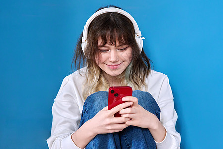 耳机中女青少年 智能手机坐在蓝色上潮人快乐工作室微笑女性技术短信电话闲暇喜悦图片