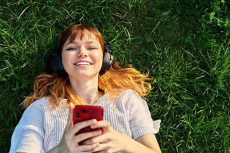 用智能手机 绿色草地的耳机充斥着红色红头发年轻女性微笑快乐播放闲暇技术电话学生女孩音乐乐趣图片