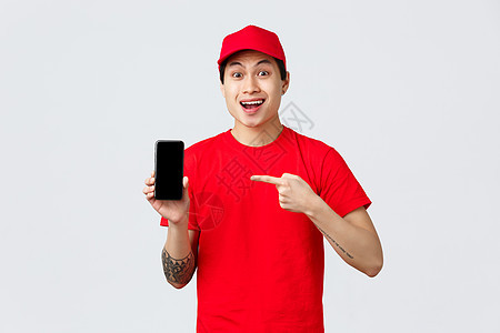 交付应用程序 在线购物和运输概念 身穿红帽和 T 恤的亚洲快递员惊讶地指着手机应用程序 展示智能手机屏幕 脸上带着有趣的表情互联图片