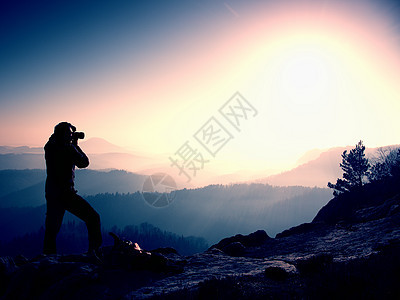 专业摄影师在岩石峰顶用镜子相机拍照 梦幻般的顽固景观 春天橙色粉红色朦胧的日出图片