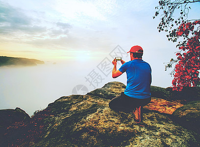 勇敢的男人坐在岩石的顶峰上 让手机照相旅游电话社会便利山脉行动自由爬坡道摄影远足图片