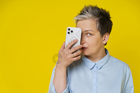 成熟的灰发女性在智能手机或现代小工具后面隐藏着害羞的微笑 工作或在线购物或查看社交媒体 黄色背景中孤立的蓝色衬衫的漂亮女人老年邮图片