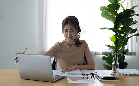 西亚亚洲女商务人士在视频电话会议上与同事团队交谈 面带微笑地在书上写笔记 使用笔记本电脑和耳机进行在线会议的女人 从家庭概念开始智能背景