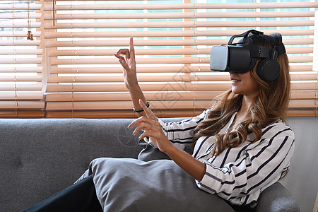 年轻女性穿着虚拟现实头盔坐在沙发上 触摸空气中的东西 未来式的生活方式是图片