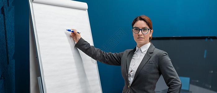 穿西装的友好女人用记号笔在空白白板上写字 红发女孩在办公室做演示 美丽的女性商务教练在一次会议上班级研讨会主持人导师教授商业推介图片