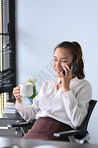 手持咖啡杯和在办公桌用手机说话的笑容女性图片