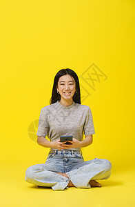 坐在地板上 手持电话的田内亚洲年轻女孩在网上发短信或购物玩游戏 孤立于黄色背景 MobileApp广告 复制空间图片