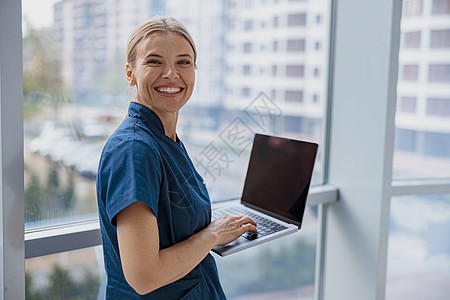 微笑的女性医护人员在医生办公室工作时使用笔记本电脑并看着相机卫生诊所医师外套技术成人桌子服务房间医院图片