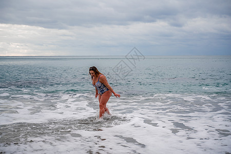 一位身穿泳衣的丰满女性在冲浪期间下水 独自在沙滩上 灰色的天空在云中 在冬天游泳海浪旅行享受海洋女孩海滩蓝色姿势孤独女士图片