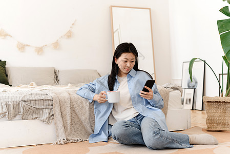 照片中一位迷人的年轻亚洲女性坐在床边的地板上 喝着咖啡 在早上享受周末 一名年轻的韩国女性一边喝着热茶 一边在手机上看新闻互联网图片