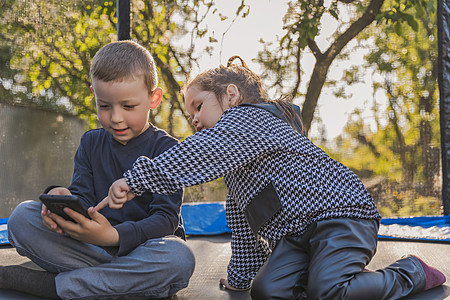 儿童坐在蹦床上看电话 他们就坐在踏板上通信儿子系统安全手机屏幕互联网娱乐影视电讯图片