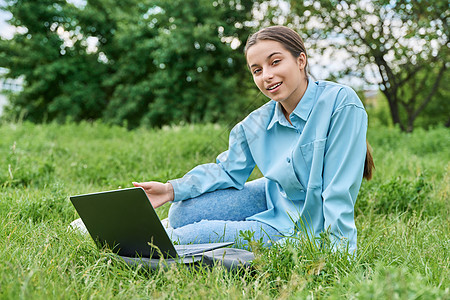 坐在绿草地上时使用笔记本电脑的高中女中学生微笑青少年青春期学习课程中学学校女士教育校园图片