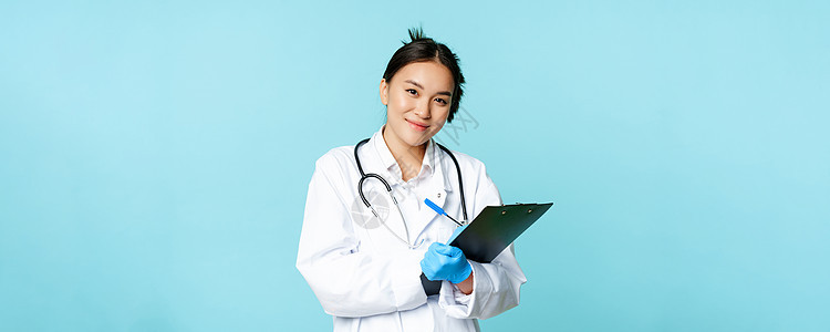 女医生的画像 身着医疗制服的亚洲医生 写下病人信息 站在蓝色背景下感染隔离预防情况疾病诊所女性护士症状考试图片