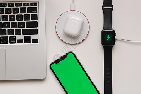 在工作场所为不同的设备充电 无线充电电缆很少 笔记本电脑 智能手表 带绿色屏幕的智能手机和白桌上的无线耳机 顶视图 小样 放置文图片