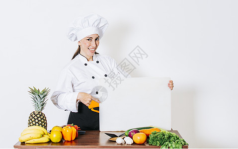 尼加拉瓜拿着空白菜单的桌上有蔬菜的厨师女人 厨房里的女厨师展示了一块空白的木板 漂亮的女厨师拿着一桌蔬菜拿着一块空白的木板营养理念女孩黑背景
