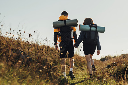 徒步旅行夫妇 年轻旅行者男女背着背包和野营垫手牵着手在路上前进的背影图片