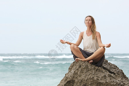 女孩坐在岩石边的海边 静坐着瑜伽女人的姿势头脑海滩闲暇训练平衡女性海岸线冥想休息女士图片