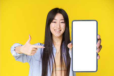迷人的亚洲女孩手持智能手机 手指在白色屏幕上 兴奋地看着黄色背景中孤立的相机 很棒的报价 移动应用广告植入式广告图片