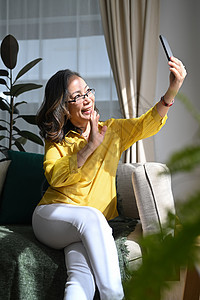 微笑的中年女士穿着休闲服 用智能手机制作 坐在家里的沙发上微笑图片