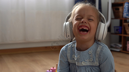 大白色耳机的孩子听的音乐 5 岁的快乐学龄前无牙小女孩在离开房间时看着相机 孩子微笑笑在家里展示舌头 童年 教育厨房房子互联网乐图片