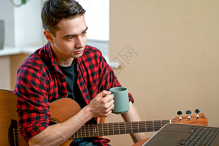 年轻人学习弹吉他 使用互联网上课 坐在咖啡桌旁 端着一杯热茶 靠近窗户的工作场所 在家工作桌子商务房间微笑成人办公室男性人士笔记图片