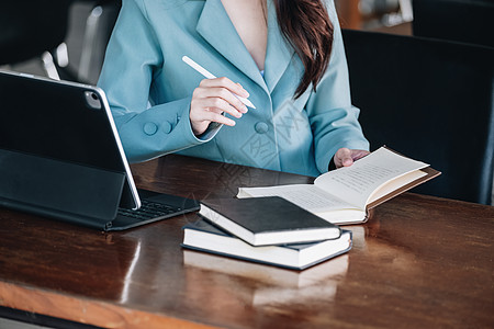 一位女企业家或女商务人士在阅读一本制定金融和投资策略的书并操作在木桌上工作的平板电脑时面带微笑商务职业笔记本员工女士同事资源战略图片