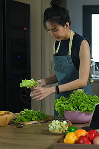 亚洲女人站在厨房桌前 准备健康的素食沙拉 (美国菜谱家)图片