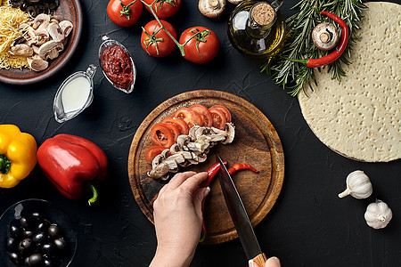 厨房桌上木板上的女性手切辣椒 蘑菇和番茄 周围是披萨的成分 蔬菜 奶酪和香料图片
