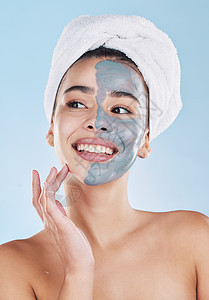 美丽的年轻混血女性在蓝色背景下应用隔离在工作室中的面罩果皮 有魅力的女人洗完澡后头上戴着毛巾 她的护肤方案让她保持新鲜淋浴身体微图片