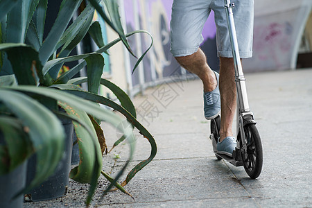 在街上穿着时髦的运动鞋骑着大轮子的城市成人滑板车的人的特写 或者在户外工作后用长剑像长龙舌兰植物一样停在公园里 看不到脸图片