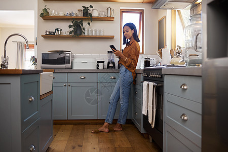 年轻的混血女人早上在厨房里一个人用手机喝着咖啡 一位西班牙裔女性在家里一边喝茶一边用手机使用社交媒体图片