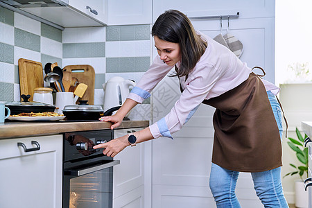年轻女子在厨房的家里烤炉里做饭成人房子烘烤厨师幸福主妇闲暇器具烤箱围裙图片