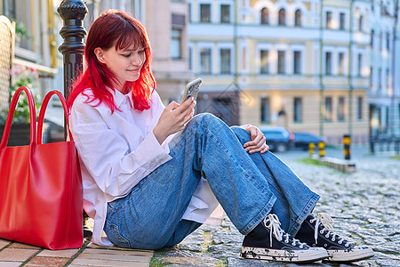 年轻时尚女性坐在人行道上 带着智能手机路面微笑衣服头发学生冒充街道红发购物者潮人图片