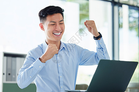 年轻快乐和兴奋的亚洲商务人士用拳头欢呼 在笔记本电脑上工作 独自坐在办公室里工作 一位中国商务人士庆祝胜利 成功后微笑的人生意人图片