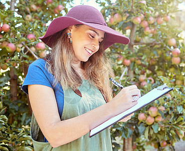 快乐的女人在阳光明媚的日子里在可持续苹果园农场工作时在剪贴板上写笔记 快乐的农民阅读清单 同时规划新鲜有机水果收获季节的任务图片