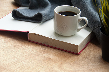 咖啡杯放在一本书的顶端 上面有灰色夹克 在木质桌边背景图片
