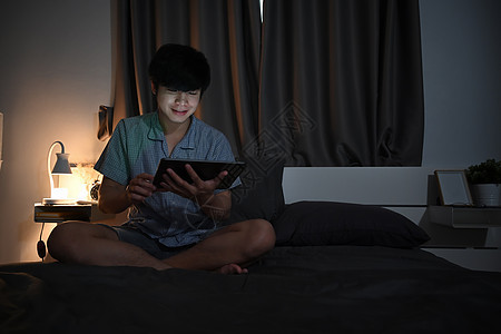 微笑的男人在晚上坐在床上时使用数字平板电脑图片