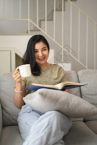 微笑的年轻女子在沙发上喝咖啡和阅读书 在家过周末休闲;图片